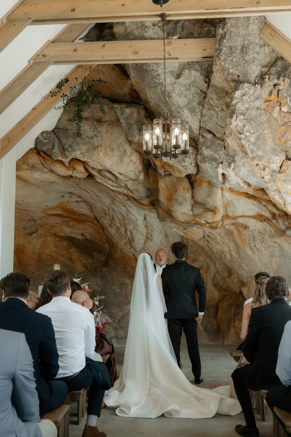 Bride in white gown and veil at cave wedding ceremony with chandelier and wooden beams.