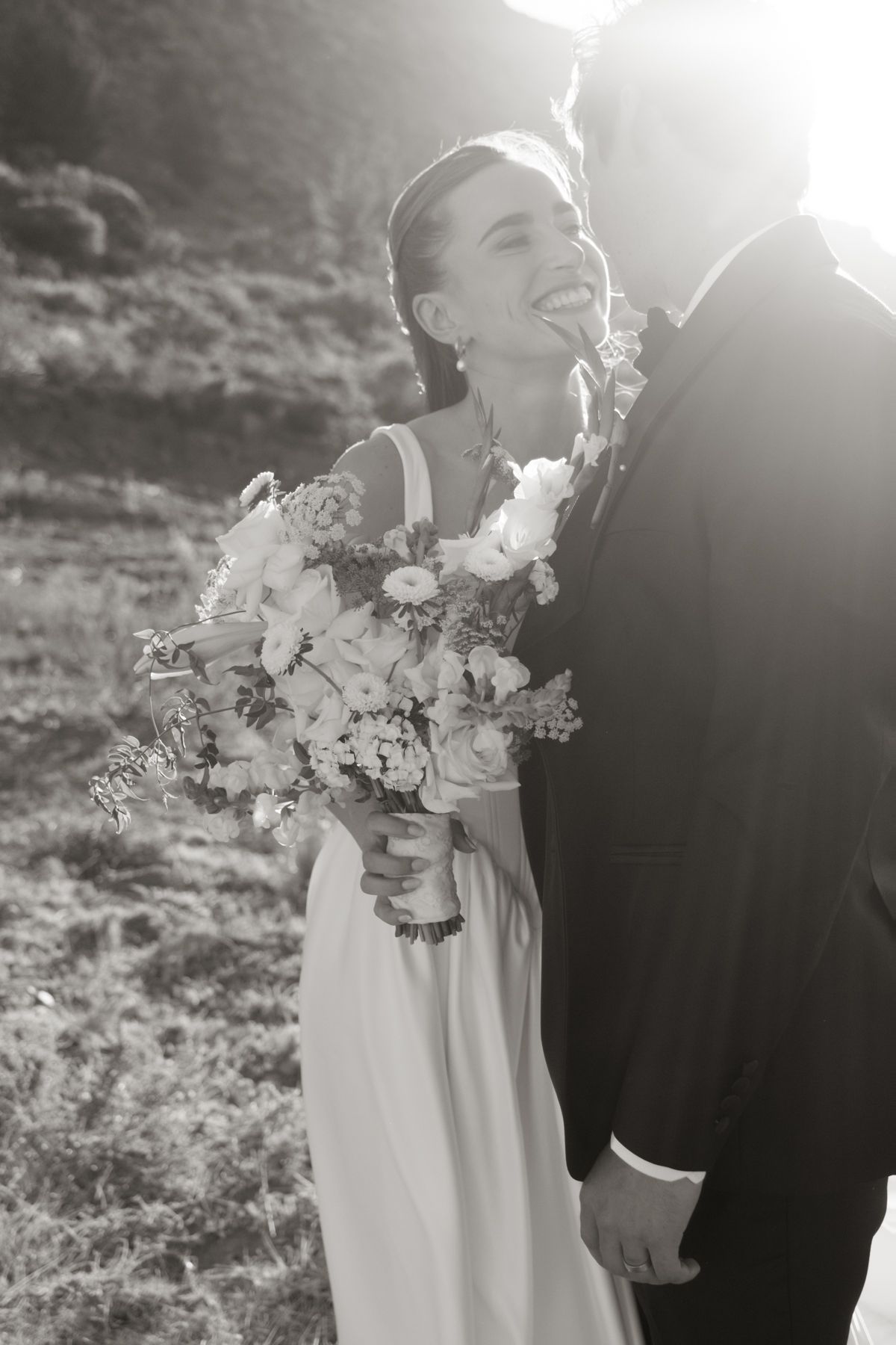 Joyful bride laughing while holding bouquet as groom nuzzles her neck in stunning black and white portrait.