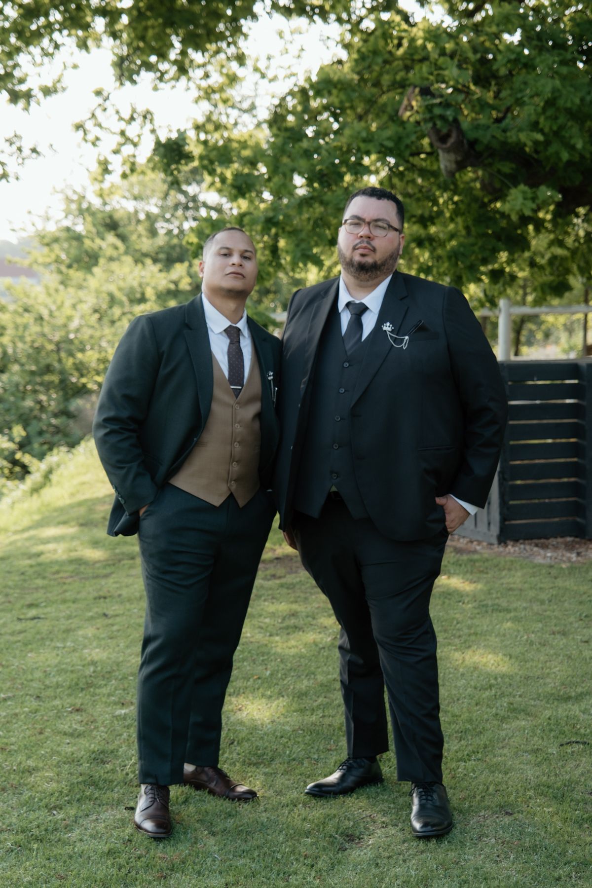 Two groomsmen in dark suits pose seriously on a green lawn at a wedding ceremony.