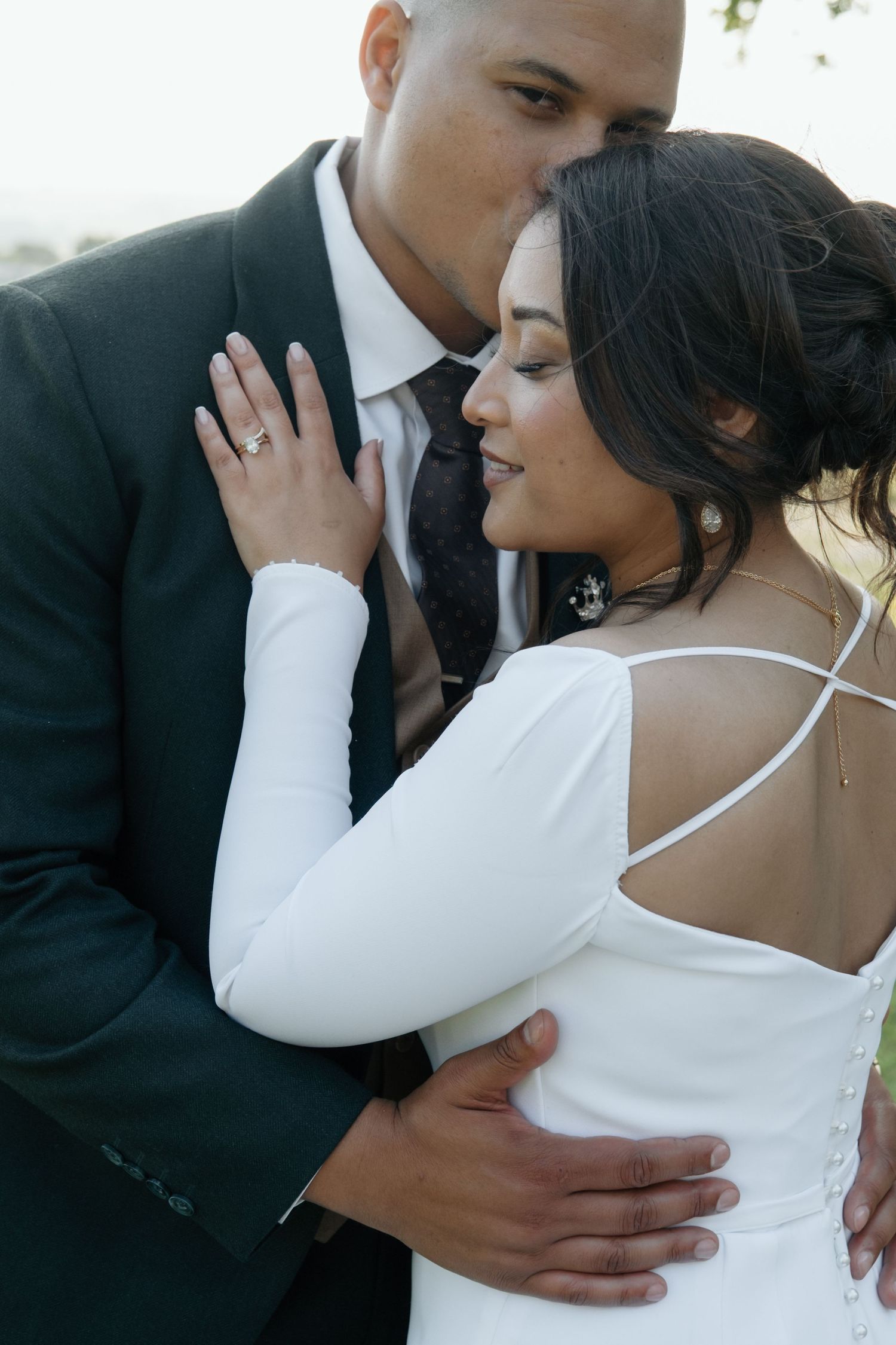 Groom in black suit kisses bride's temple while embracing her in elegant white long-sleeve wedding gown outdoors.