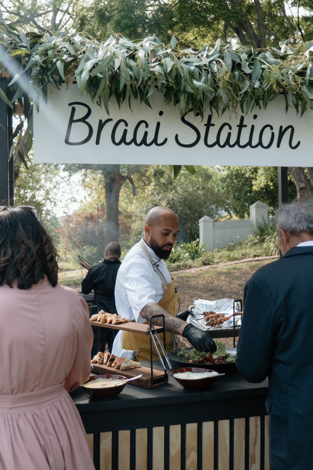 Chef serving guests at an outdoor Braai Station food stall decorated with eucalyptus garland.