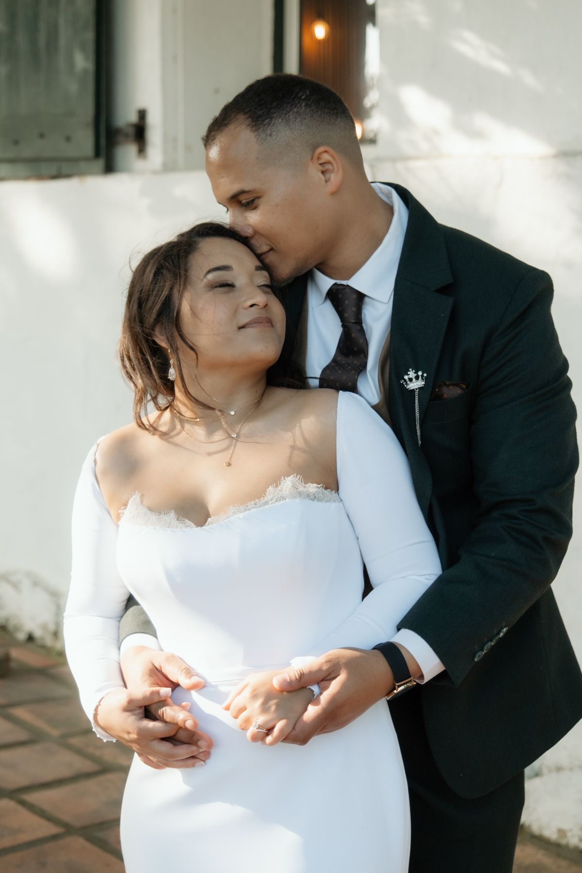 Groom in dark suit tenderly kisses bride on forehead while embracing her against white wall.