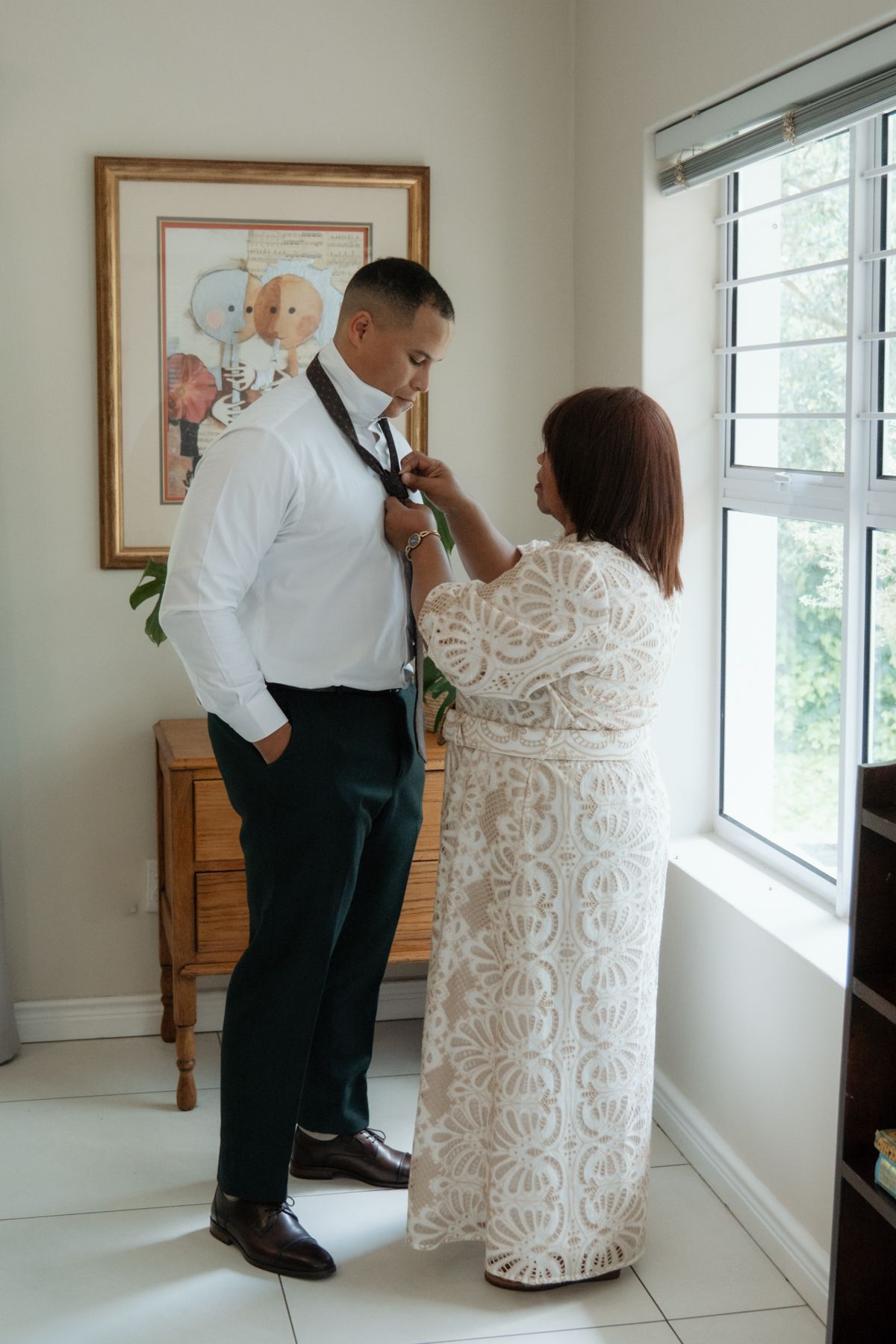 Woman in lace dress adjusting groom's boutonniere near a bright window during wedding day preparations indoors.