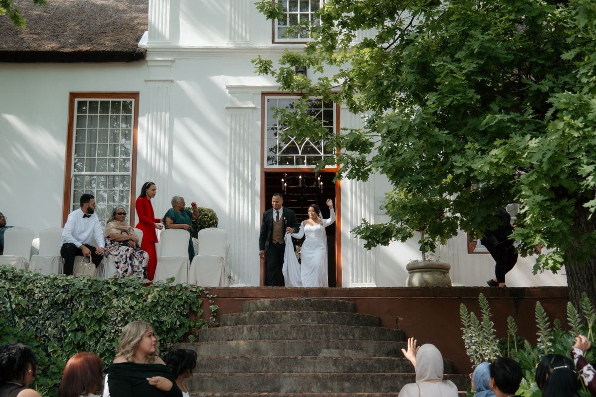 Bride and groom emerging from historic green manor house doorway to waiting wedding guests.