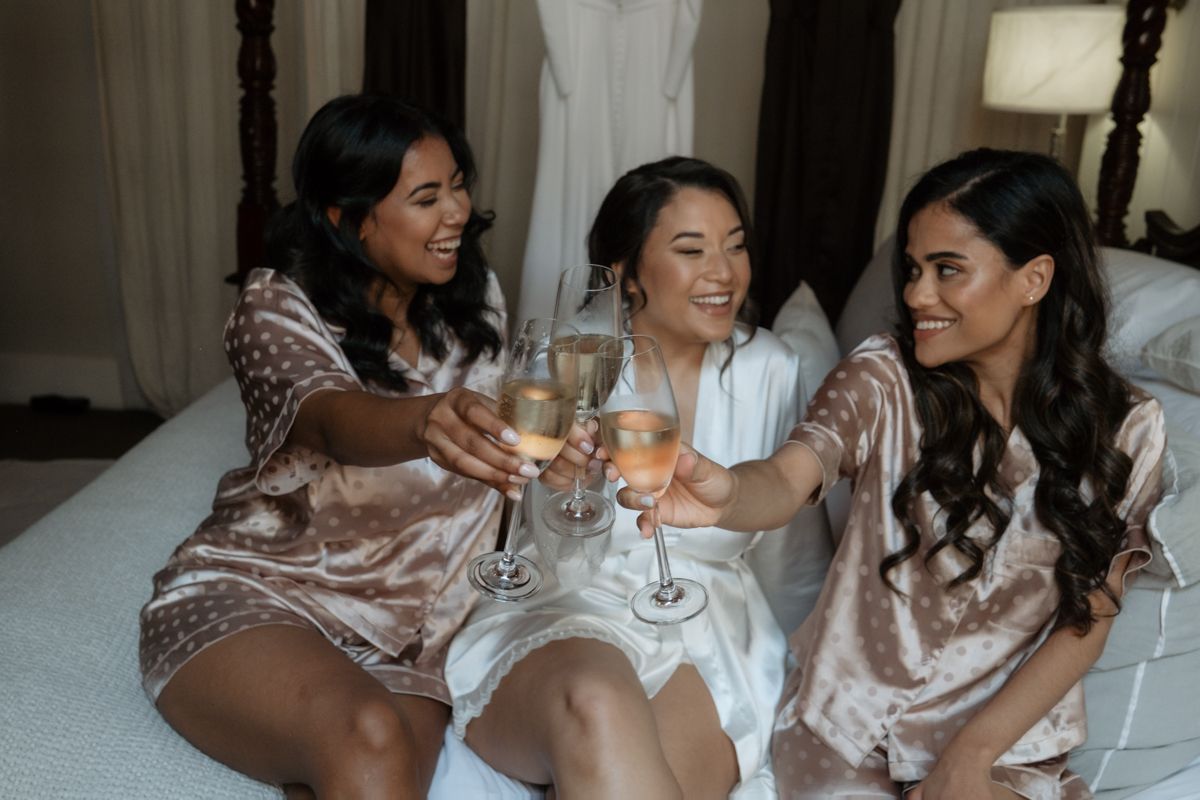 Bride in white robe toasting champagne with two bridesmaids in satin robes on a bed.