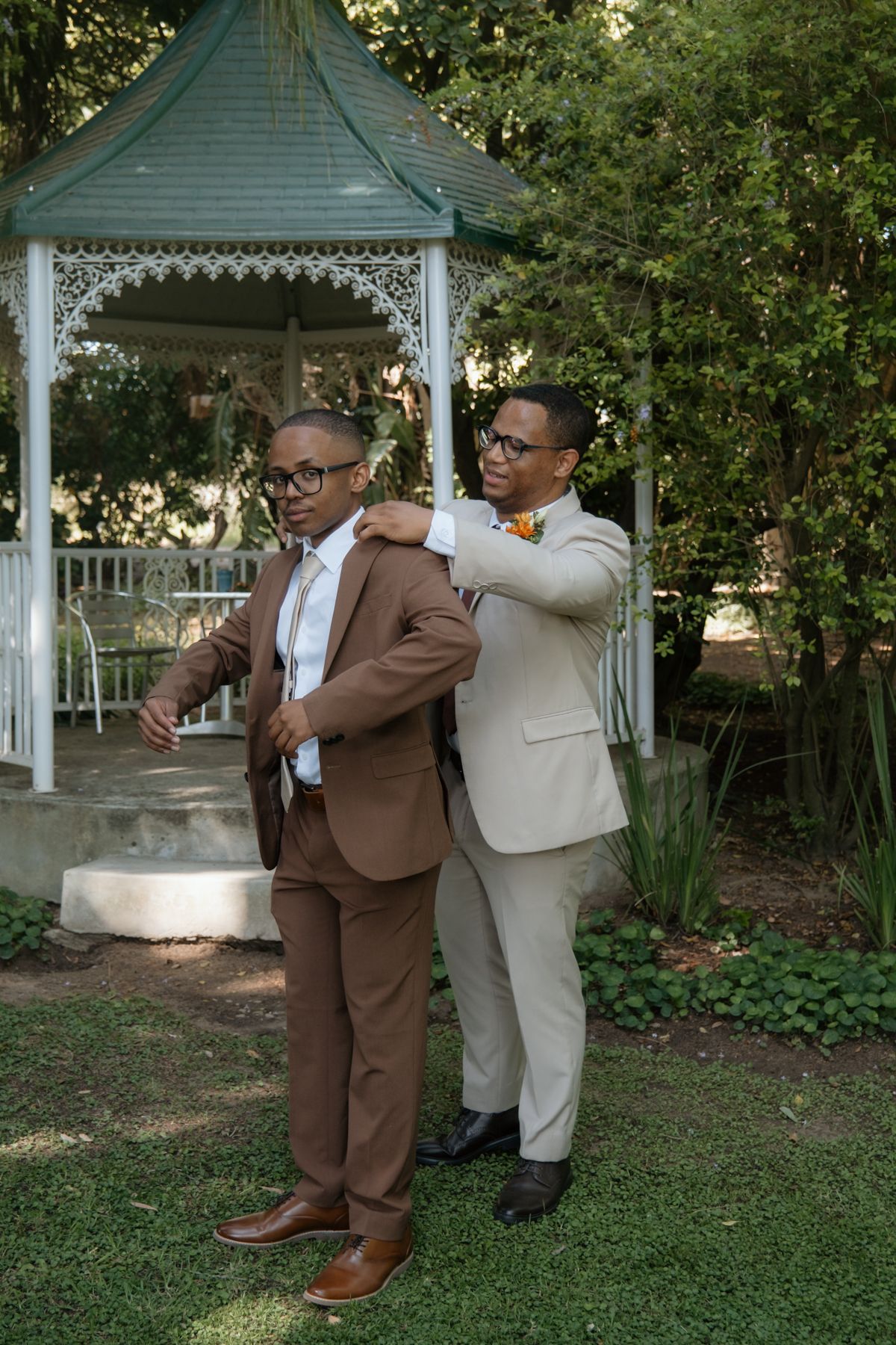 Groom in brown suit gets jacket adjusted by groomsman in beige suit near garden gazebo.