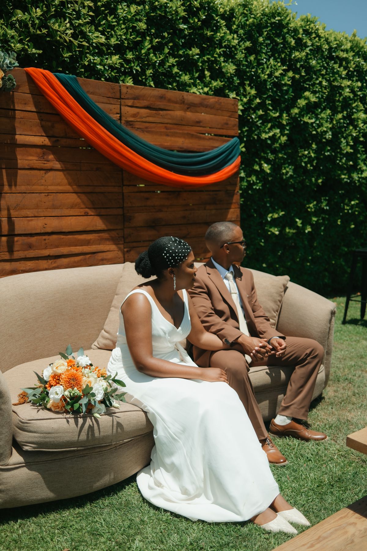 Bride and groom sitting on beige outdoor sofa with orange bouquet, wooden backdrop draped with orange and teal fabric.