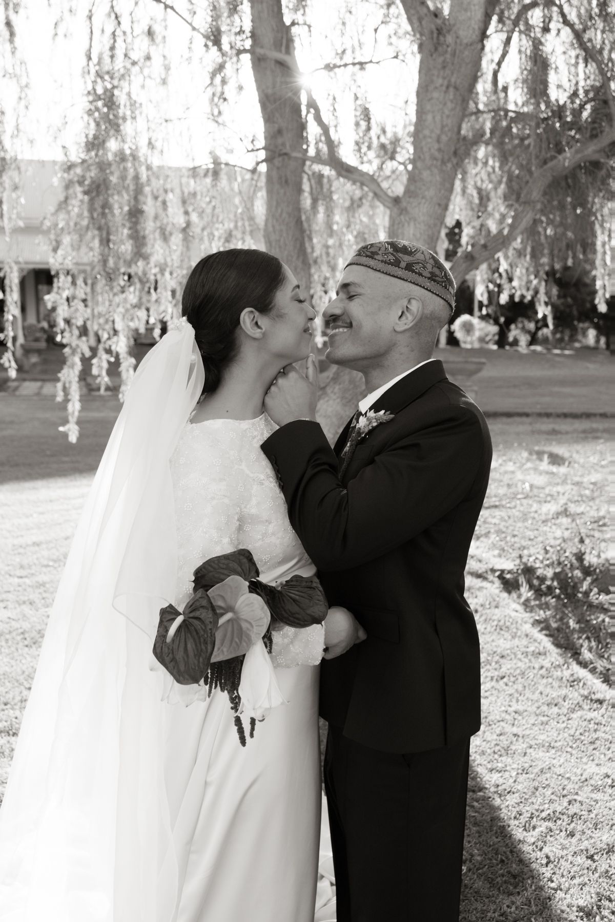 Black and white photo of bride kissing groom on cheek under a weeping willow tree at wedding.