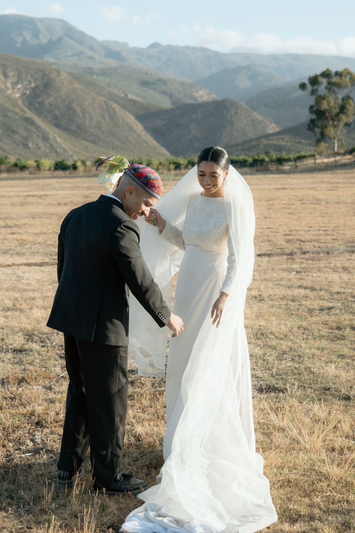Groom in colorful kufi adjusts brides veil as she laughs in sun-drenched mountain wedding field.