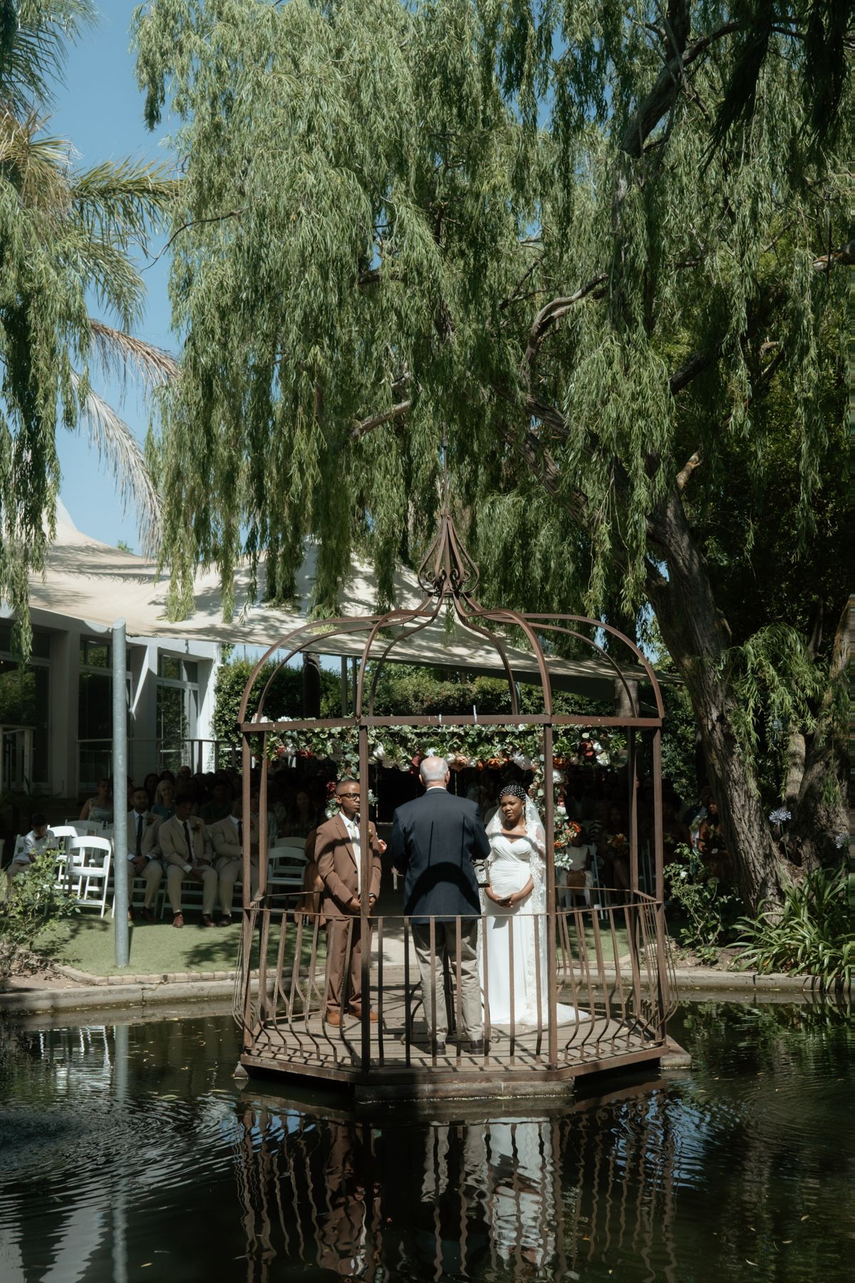 Wedding ceremony on ornate gazebo island surrounded by pond under willow trees.