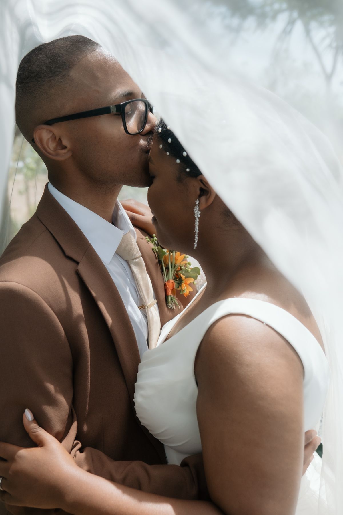 Newlyweds sharing intimate kiss under bridal veil, groom in brown suit with glasses, bride in white sleeveless wedding dress.
