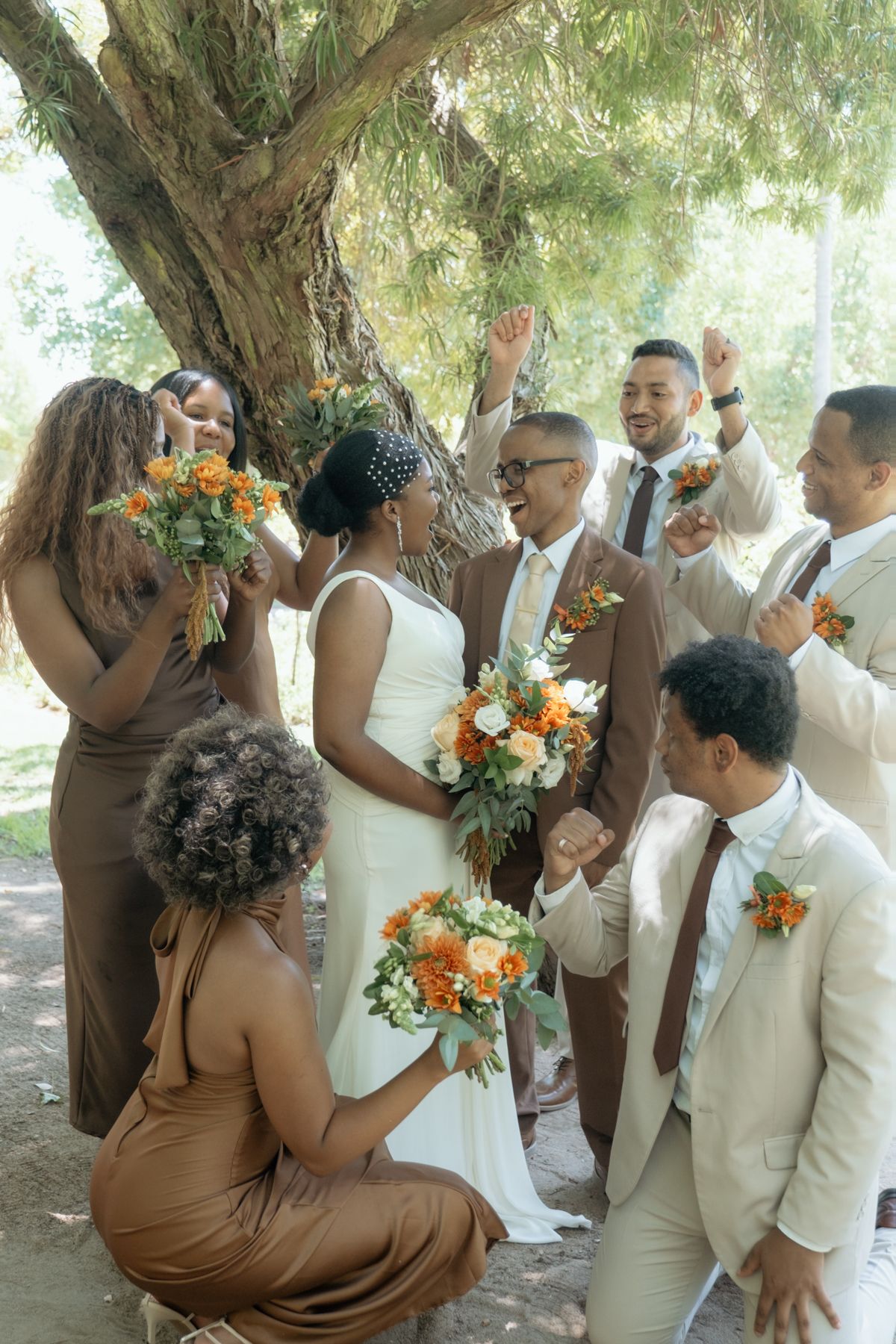 Joyful wedding party celebrates under a tree, bridesmaids in brown, groomsmen in tan suits, bride holds orange floral bouquet.