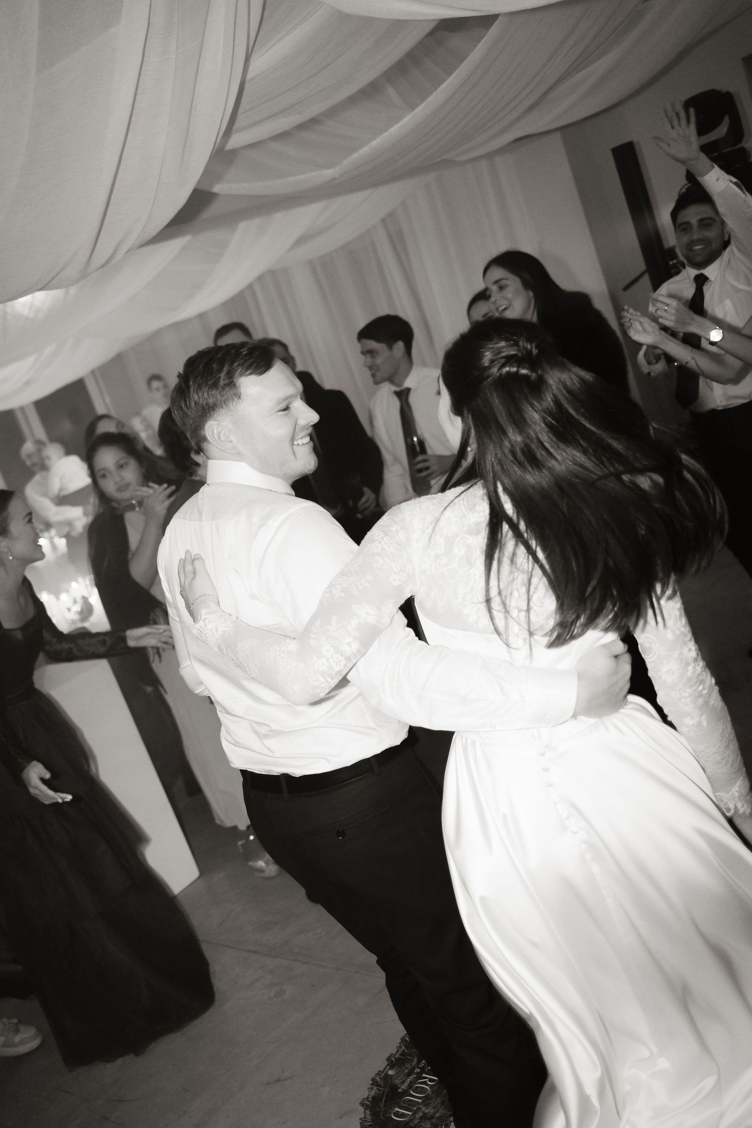 Black and white photo of joyful bride and groom dancing energetically with guests at wedding reception.