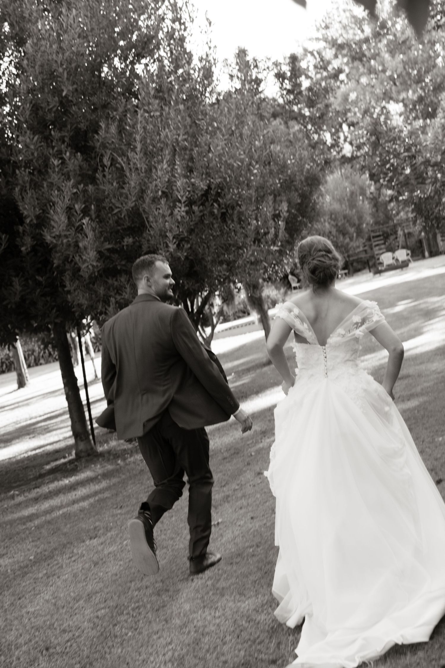 Black and white photo of bride and groom walking hand in hand through a tree-lined garden.