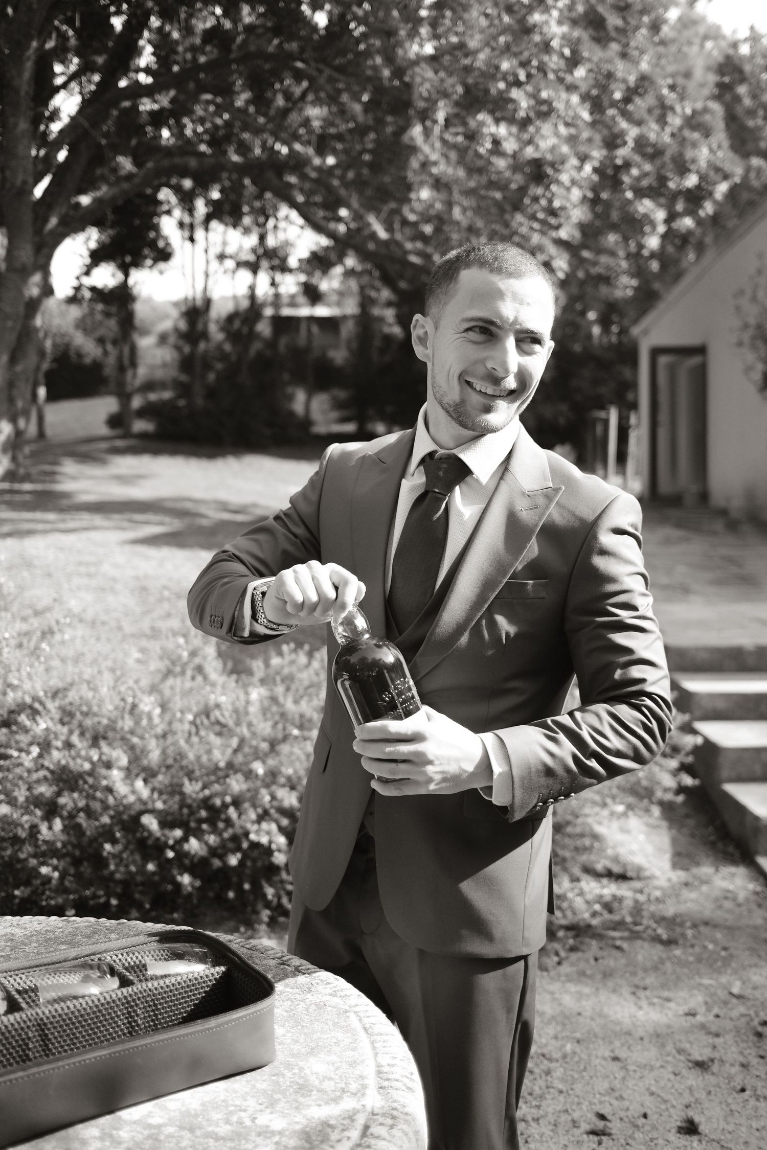 Smiling groom in gray suit opening champagne bottle outdoors at wedding venue in black and white photo.