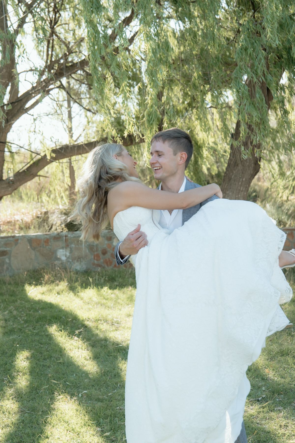 Groom lifting smiling bride in white gown outdoors under a green willow tree.