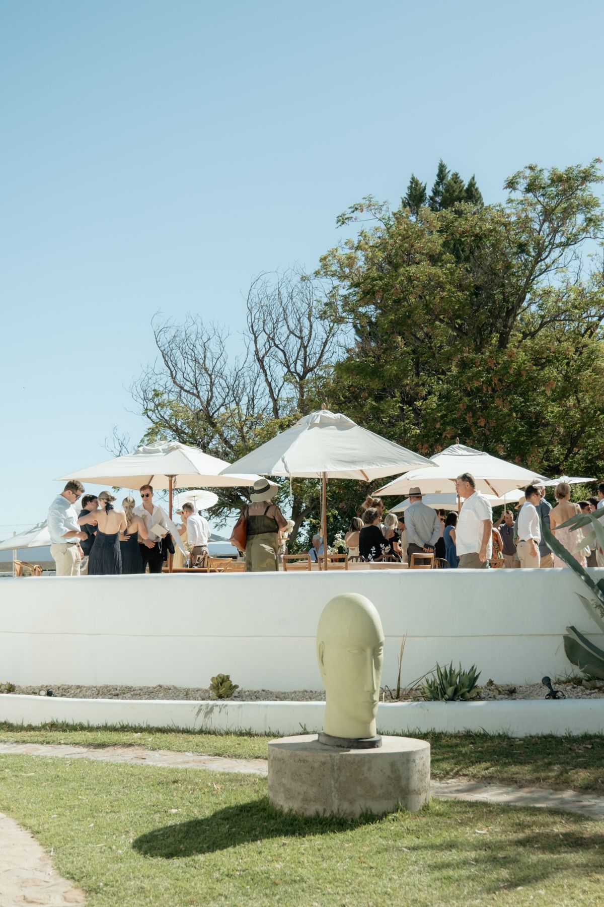 Wedding guests mingling under white umbrellas on a sunny outdoor terrace with trees.