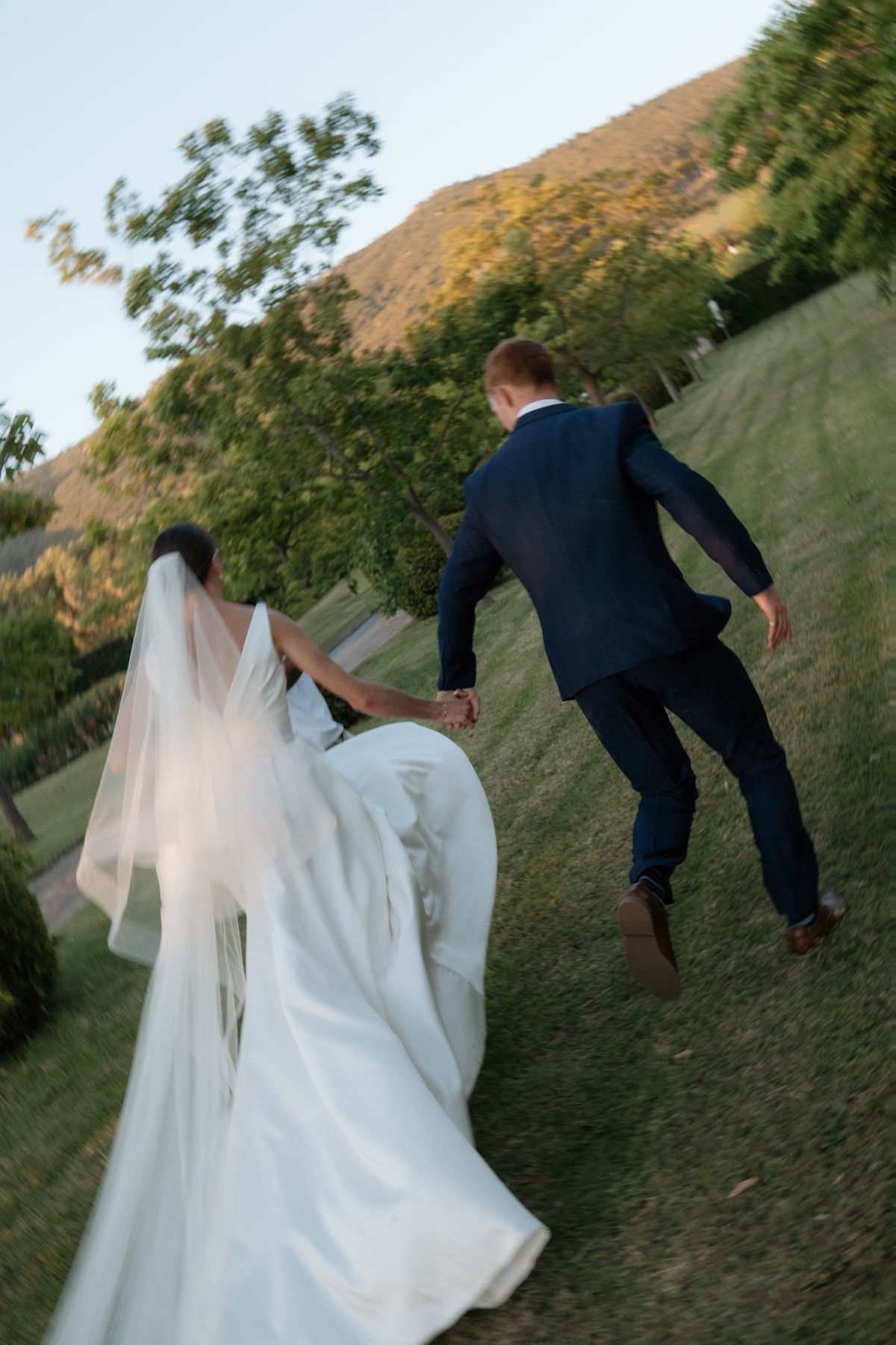 Bride and groom holding hands while running up a grassy hill with scenic mountains in the golden evening light.