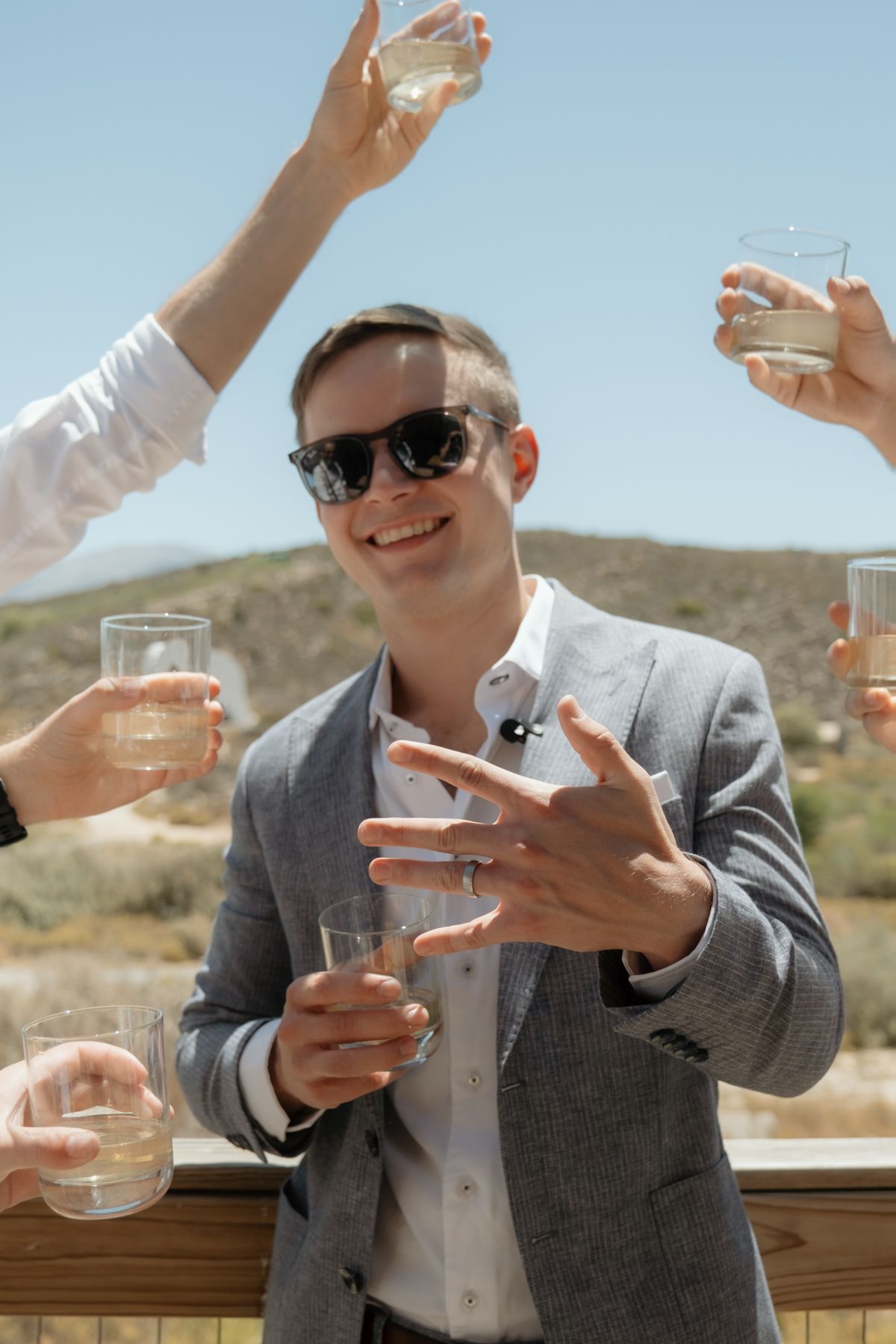 Smiling man in gray blazer and sunglasses raises a glass during an outdoor toast with friends on a sunny day.