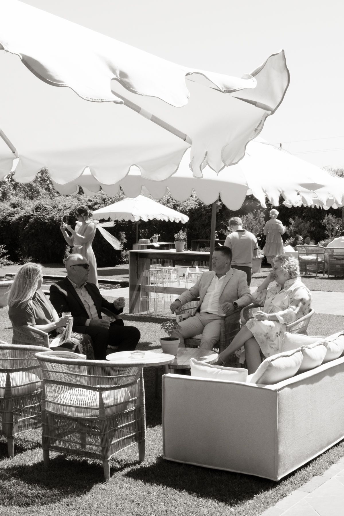 Wedding guests relaxing on outdoor lounge furniture under white parasols at sunny garden reception.