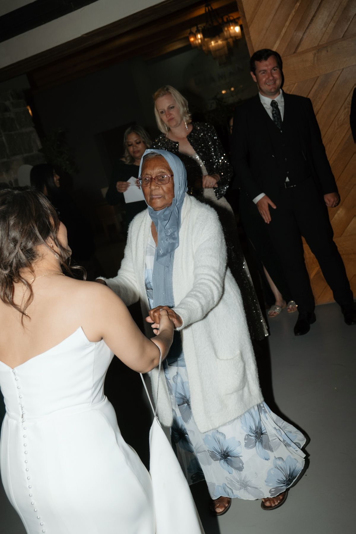 Bride in white gown holds hands and dances with elderly guest in floral dress at wedding reception.