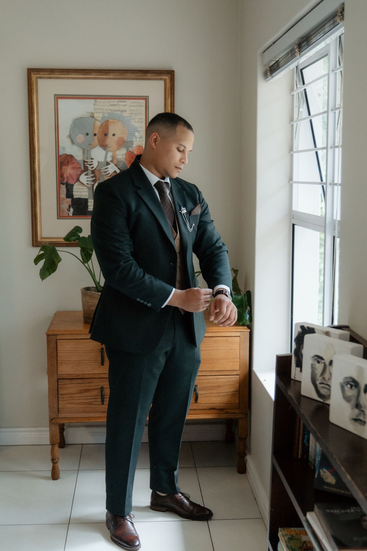 Dapper groom in dark green suit adjusts cufflinks while standing by window before wedding ceremony.
