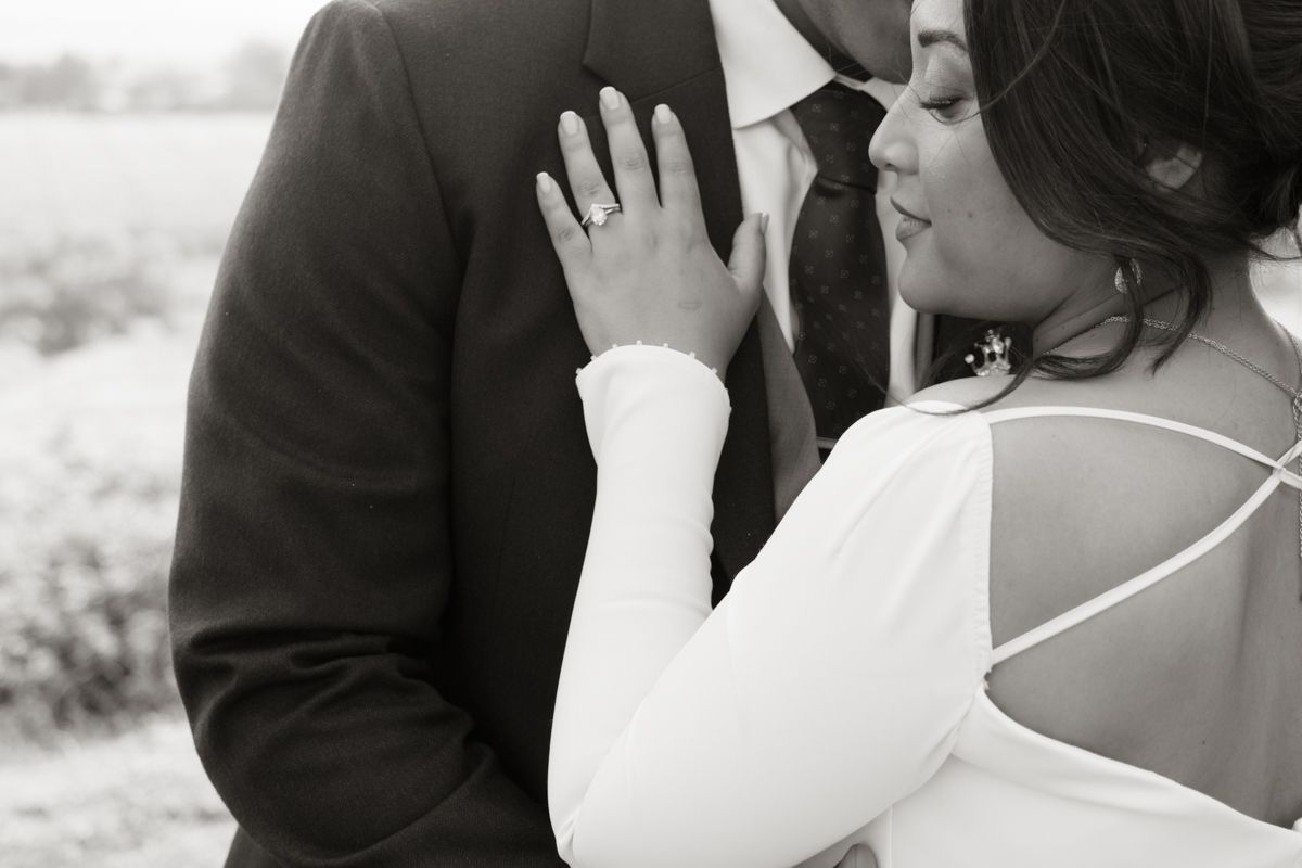 A groom kisses his bride tenderly while she displays her engagement ring in black and white.