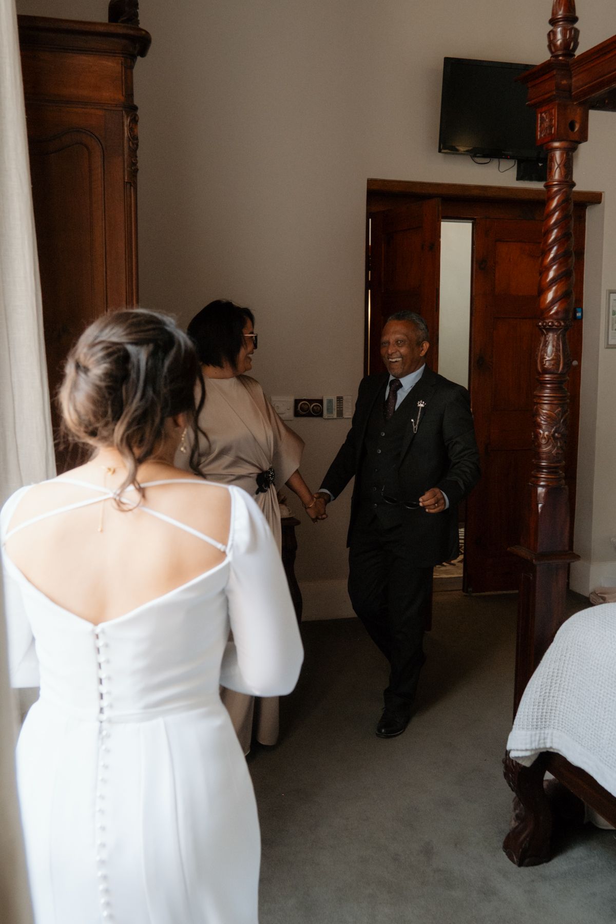 Bride in white gown with cross-back detail greets elderly family member entering a elegant wood-furnished bridal suite.