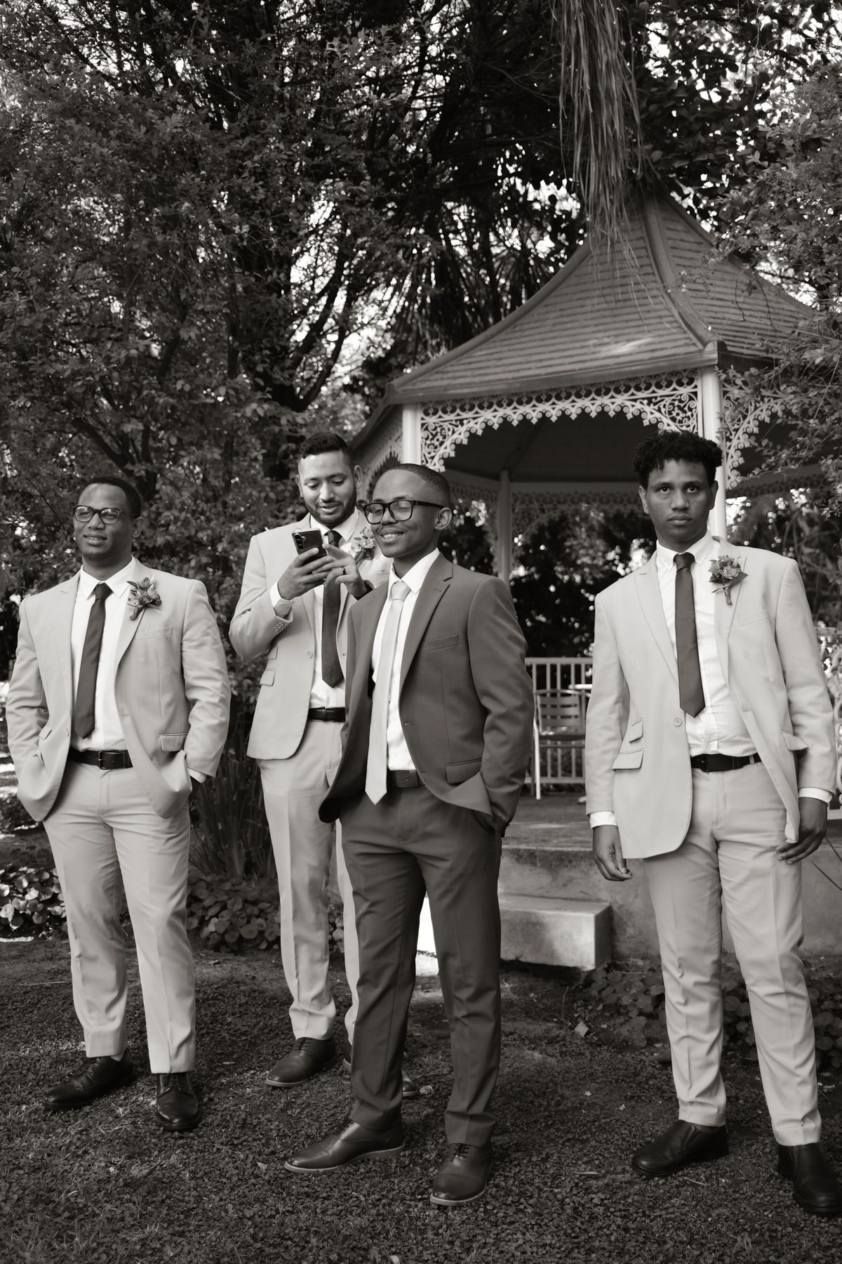 Black and white photo of four groomsmen in suits relaxing near decorative garden gazebo.