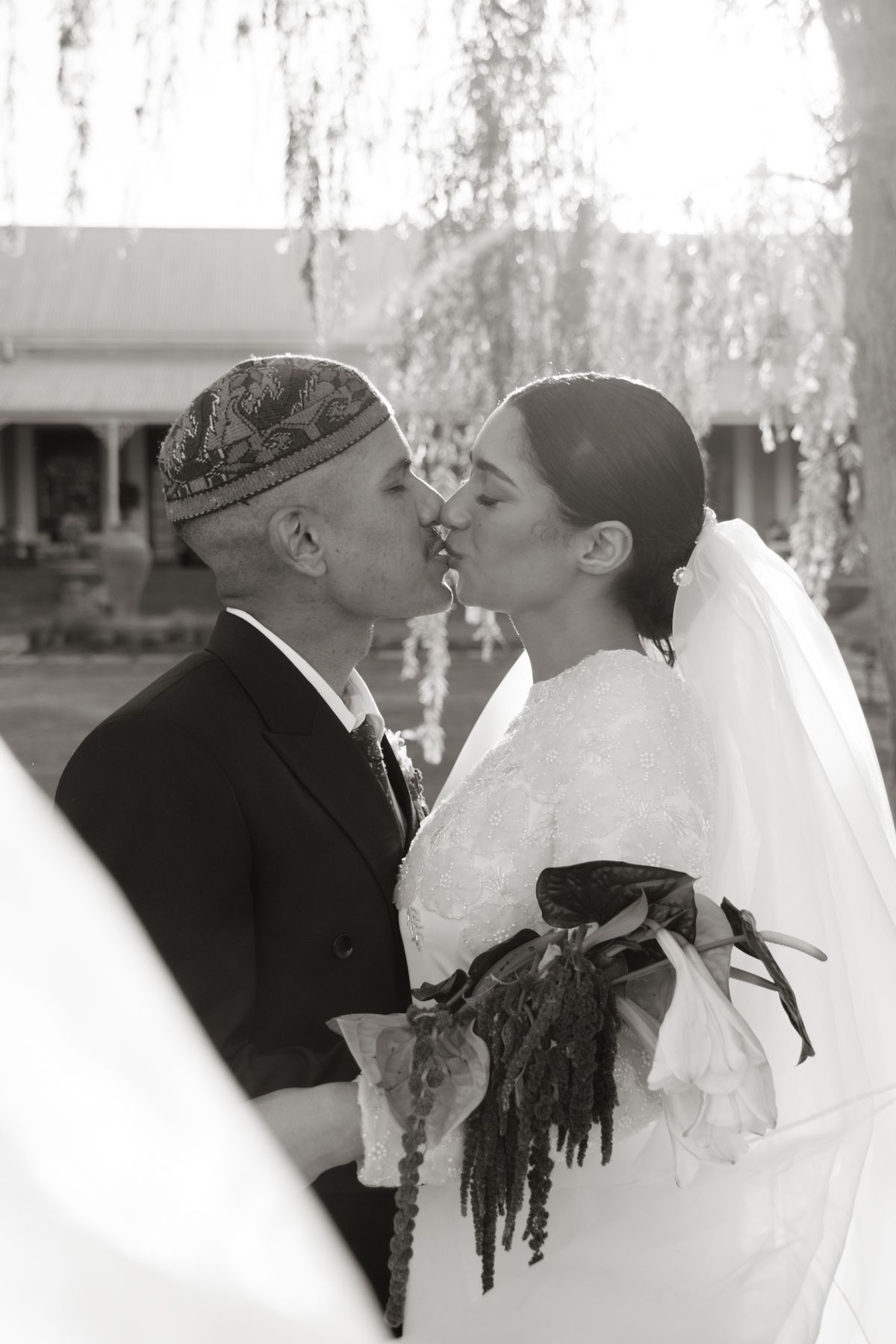 Black and white photo of bride and groom sharing a kiss under a willow tree.