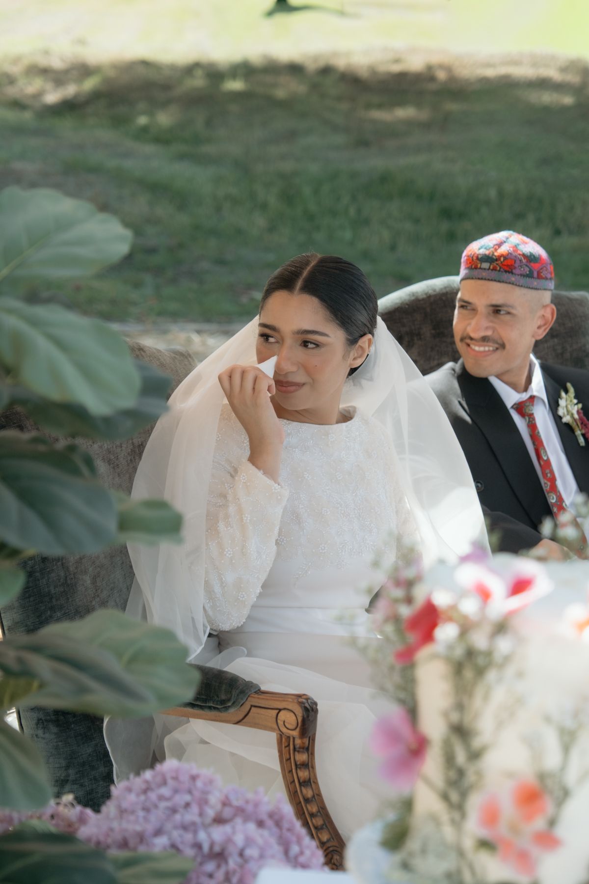 Emotional bride in white veil wipes tear while groom in embroidered kippah smiles at outdoor wedding.