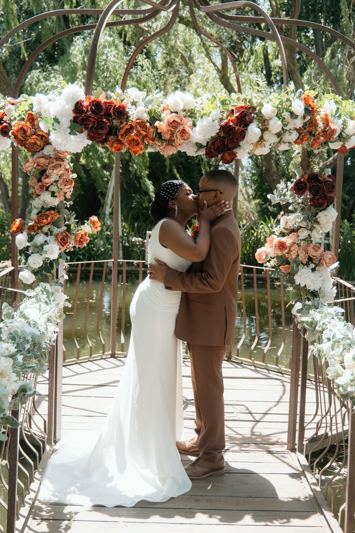 A Black couple shares their first kiss as newlyweds under a floral arch adorned with orange, red, and white roses outdoors.