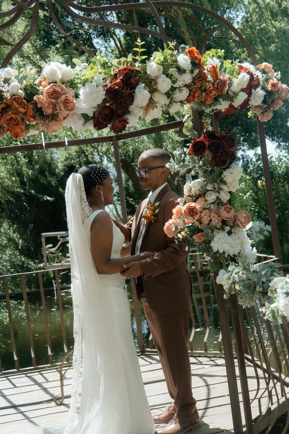 Bride and groom exchange vows under floral arch with orange, red, and white roses at outdoor wedding.