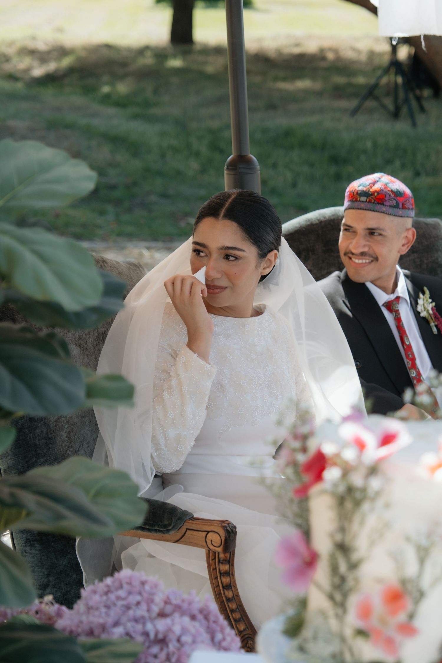 Bride in white lace gown and veil sits beside smiling groom wearing colorful kippah at outdoor wedding reception.