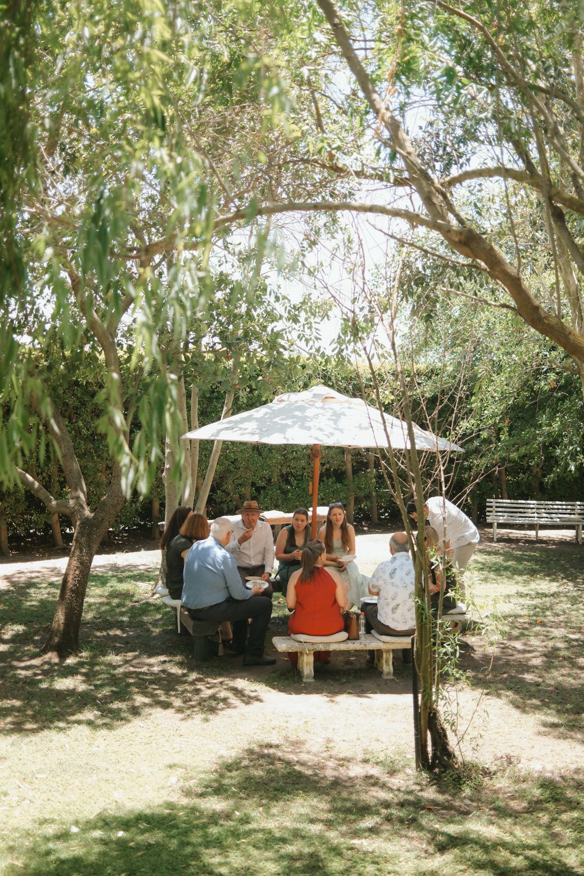 Group of people gathered at an outdoor picnic table under a large umbrella surrounded by lush green trees on a sunny day.