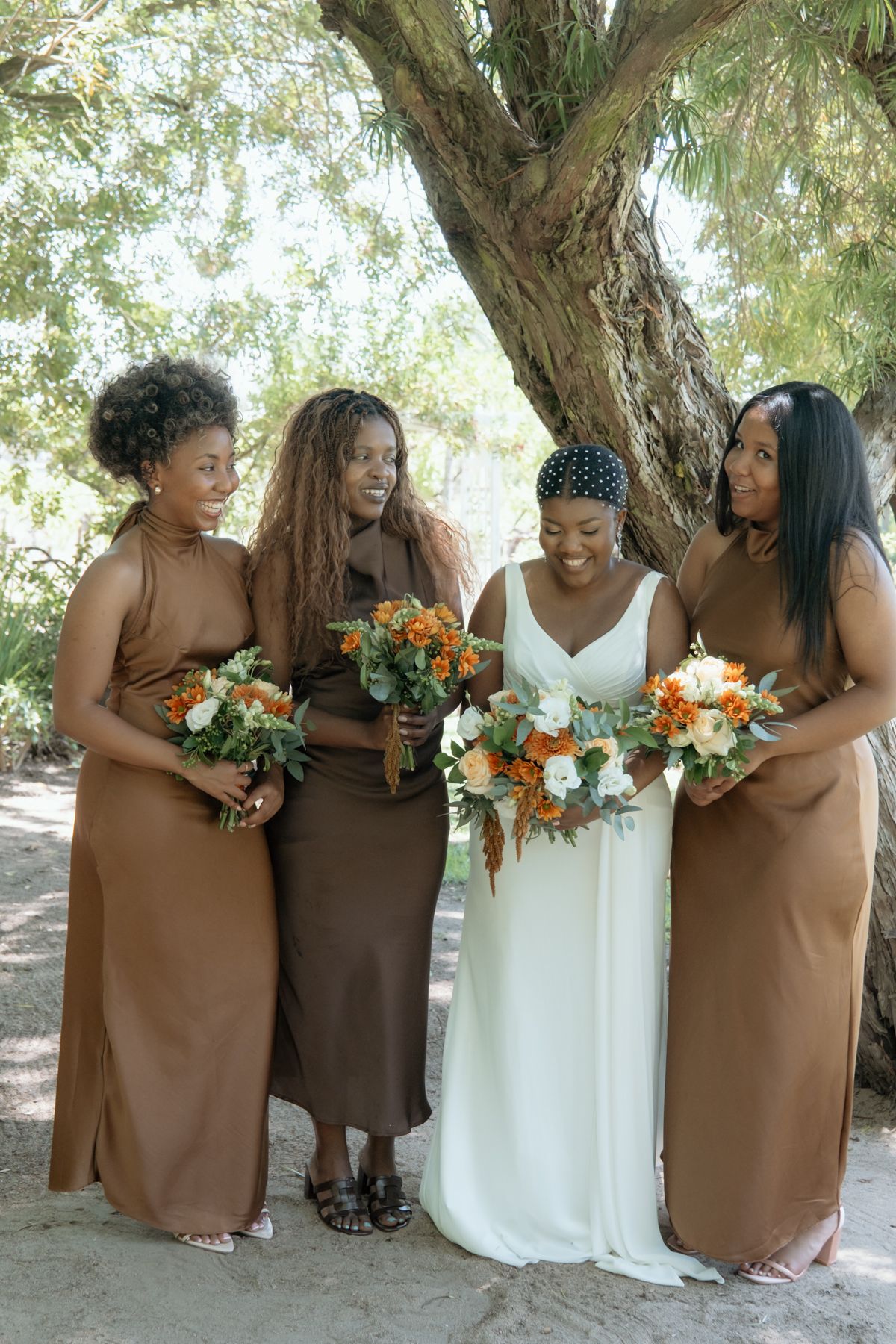 Bride in white gown stands with three bridesmaids in brown dresses holding orange and white bouquets under a shady tree.