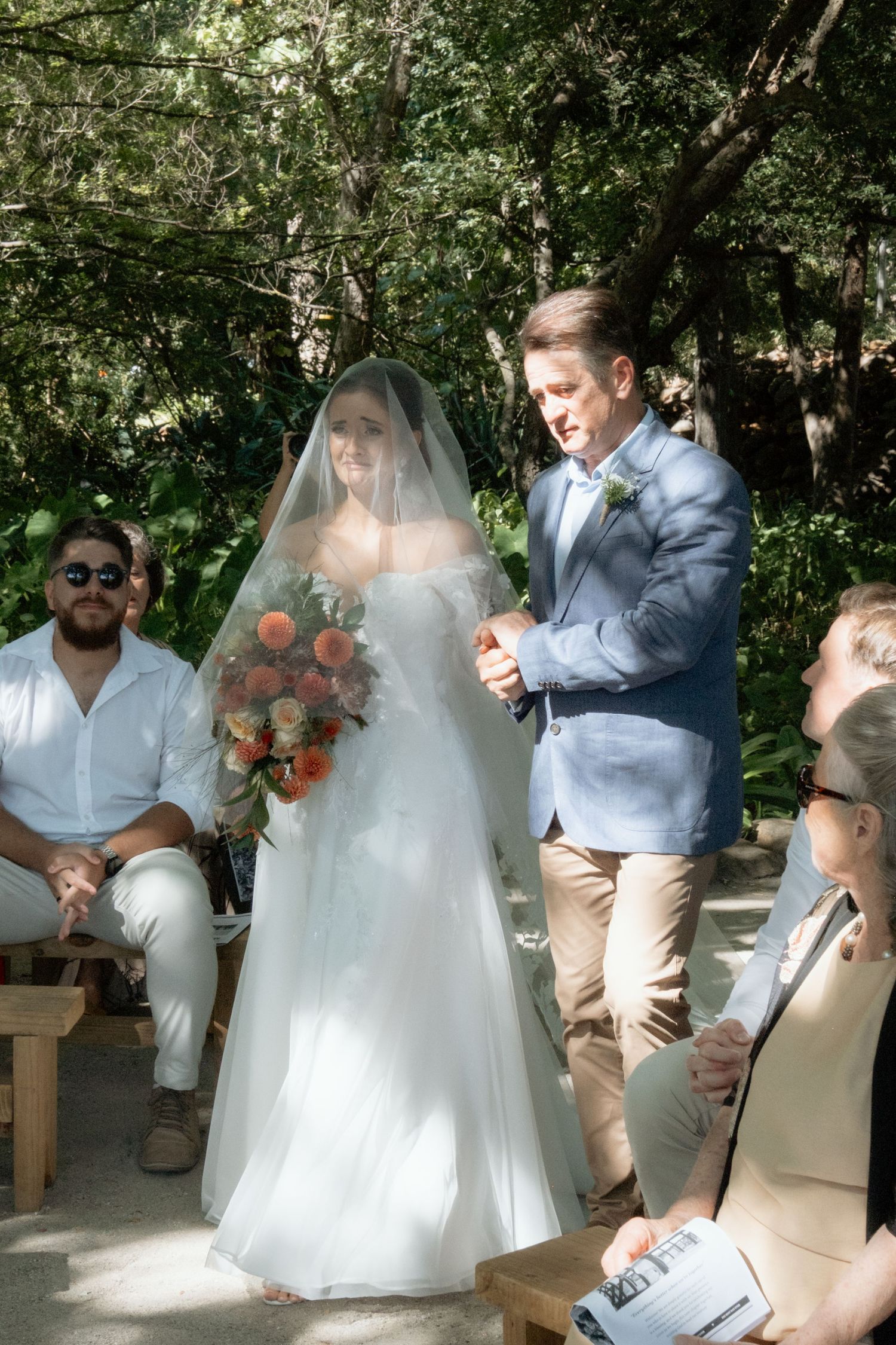 Bride in white gown with bouquet walking down the aisle with escort at outdoor garden ceremony.