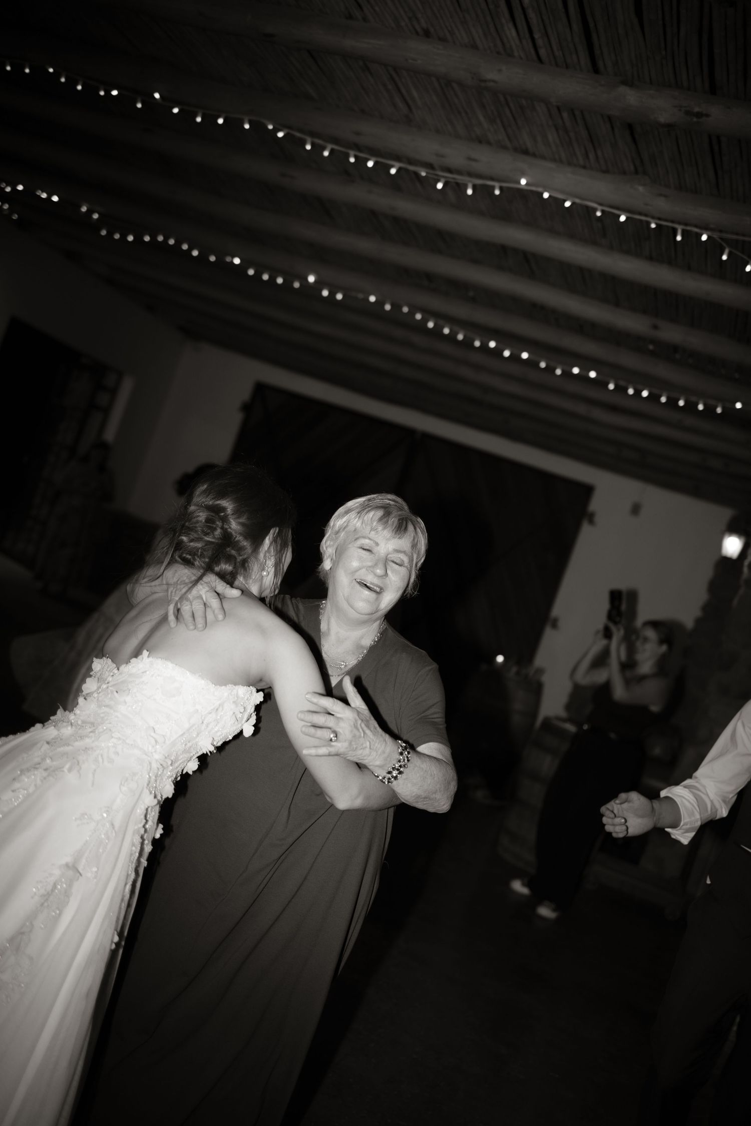 Black and white photo of bride dancing joyfully with older woman under fairy lights at wedding reception.