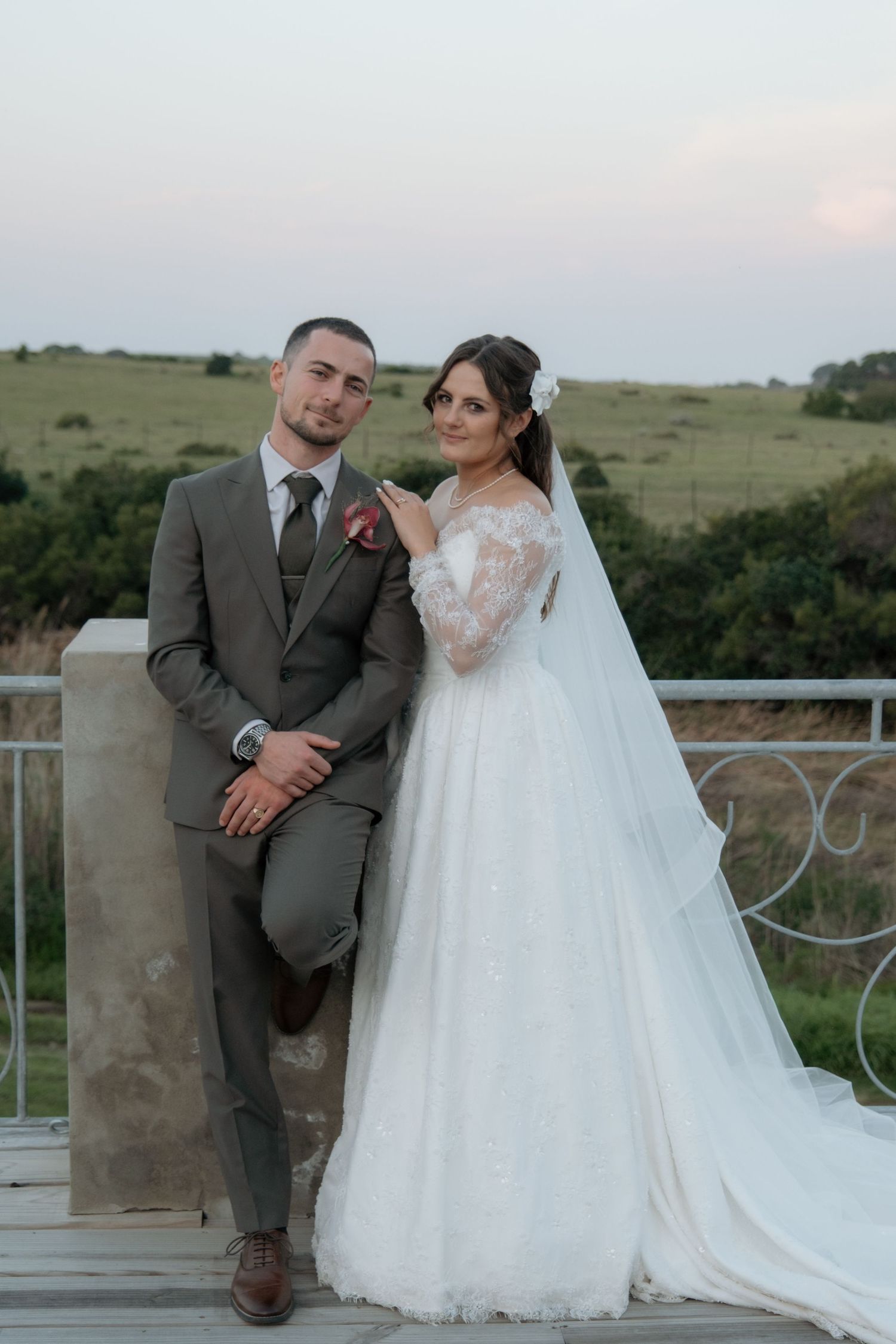 A bride in a white lace gown and groom in a gray suit pose together on a balcony with green fields at sunset.