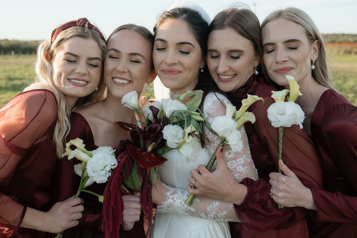 Bride in white gown with four bridesmaids in burgundy dresses holding white and red bouquets outdoors.