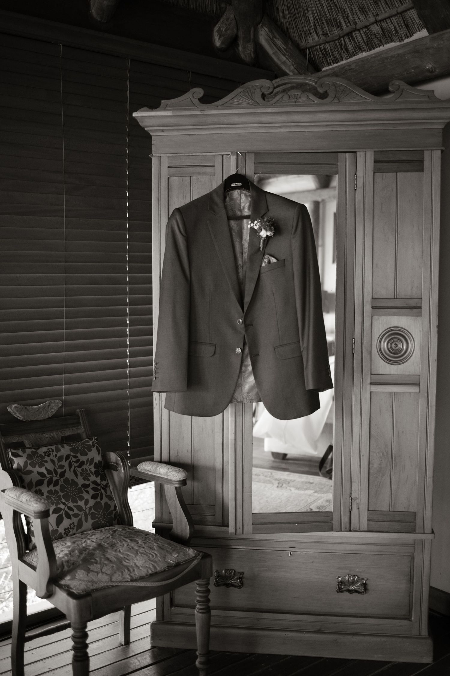 Black and white photo of a groom's suit jacket with boutonniere hanging on a vintage wooden armoire.