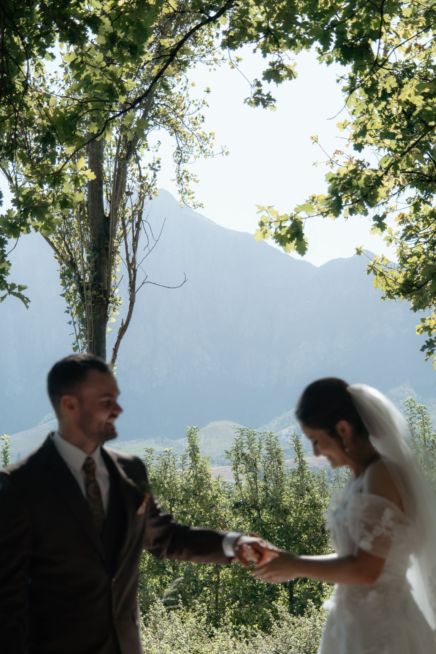 Smiling groom holds bride's hands under green trees with dramatic misty mountains in the background on wedding day.