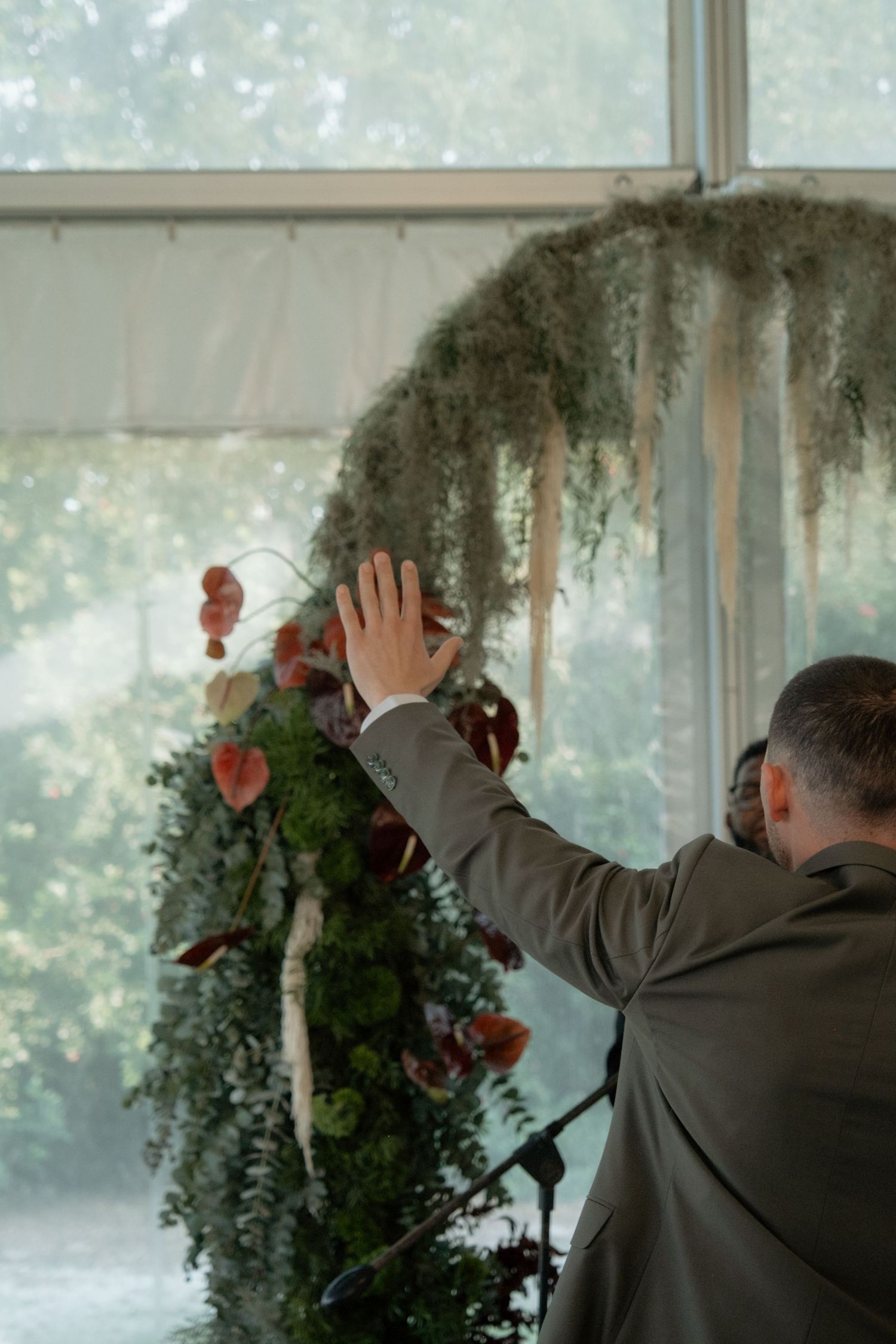 Groom in olive suit adjusts lush floral arch with greenery and red flowers at wedding ceremony.