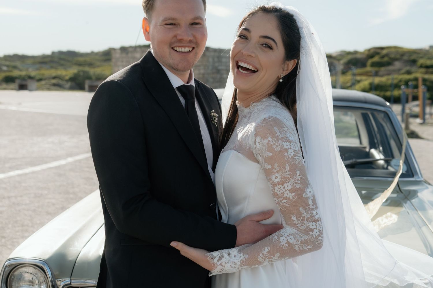 Happy bride and groom laughing together in wedding attire beside a vintage car outdoors on their wedding day.