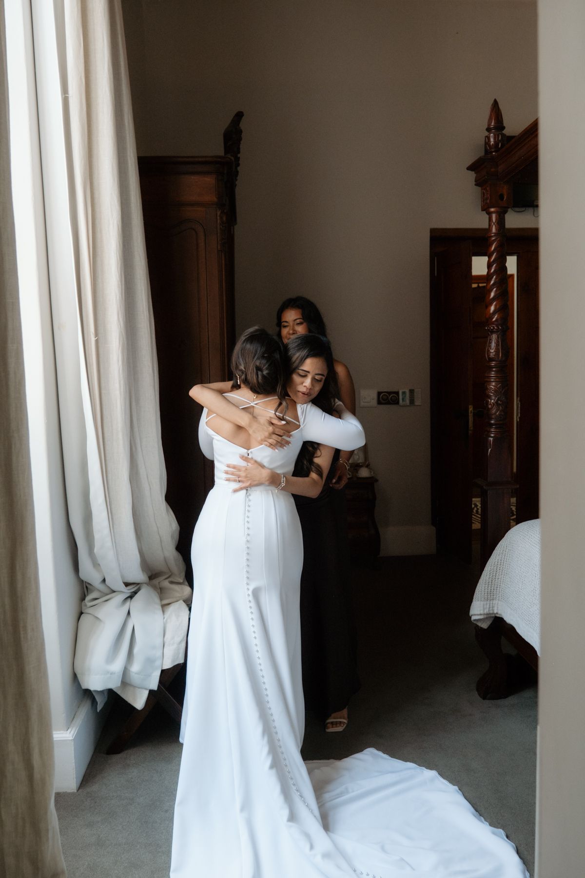 Bride in fitted white gown with button train shares emotional hug with loved one in elegant bridal suite.