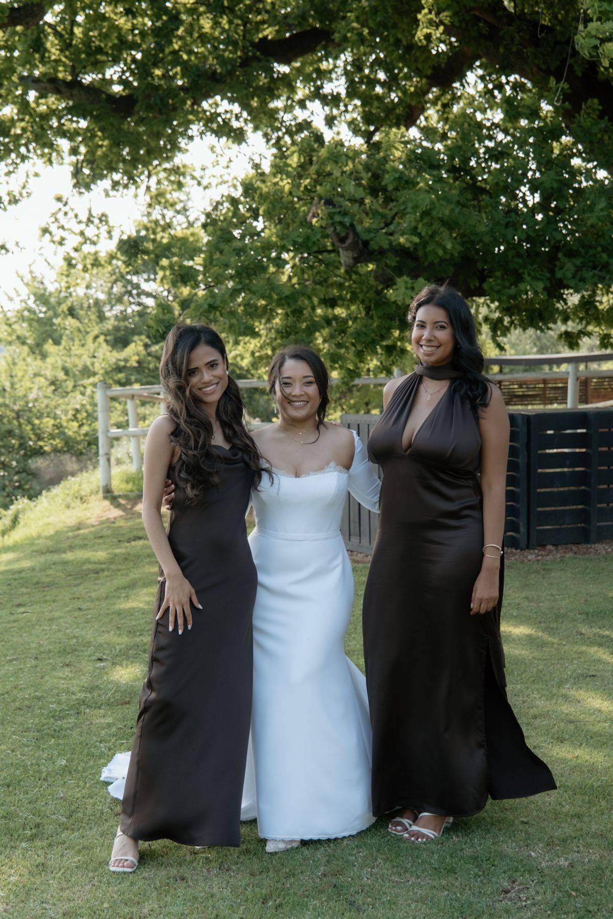 Bride in white gown smiles with two bridesmaids wearing elegant brown satin dresses outdoors under a green tree.