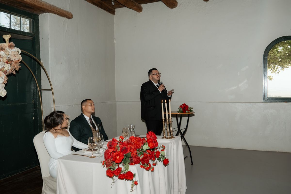 Wedding guest delivers a heartfelt speech at the reception dinner table adorned with red floral arrangements.