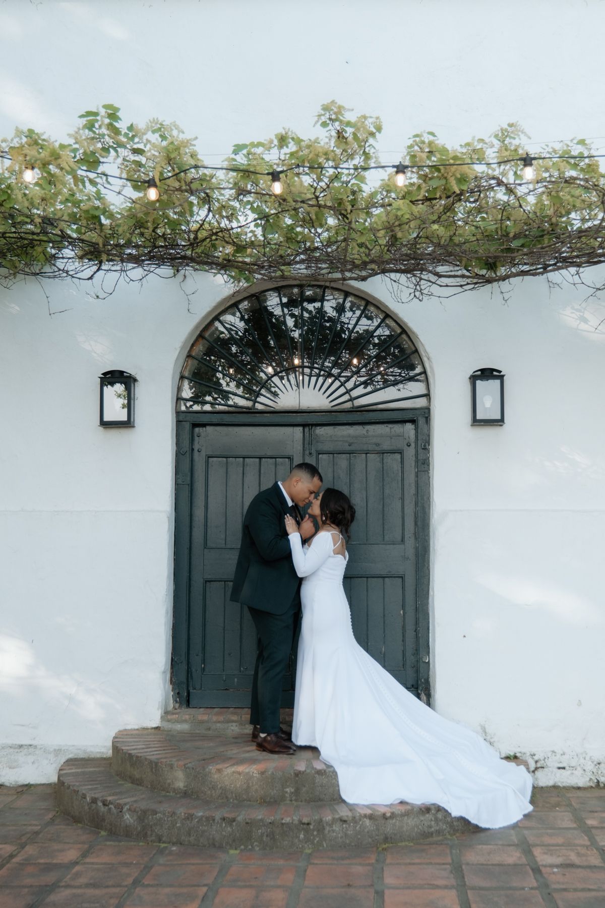 Bride and groom share a romantic kiss at an arched wooden door adorned with fairy lights and vines.