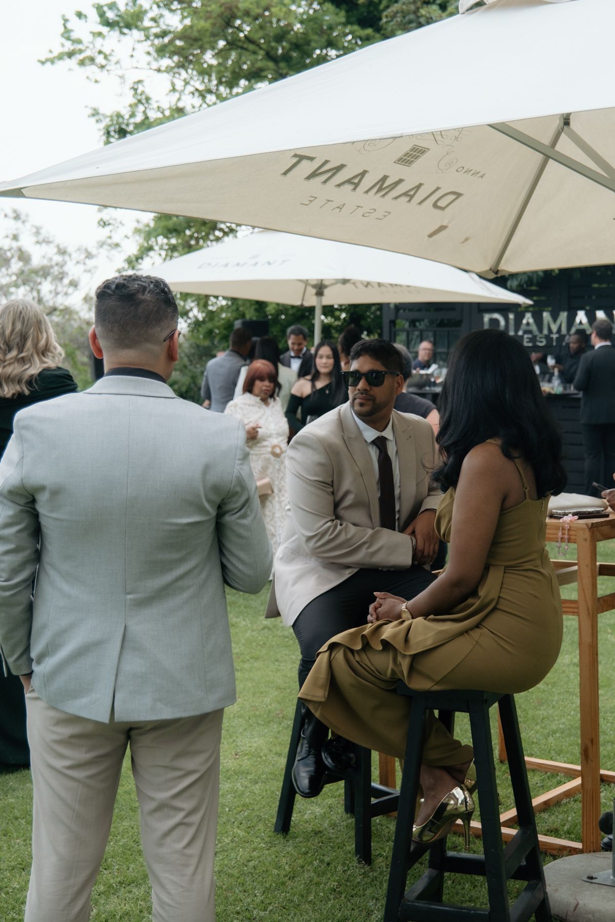 Wedding guests socializing under Diamant Estate umbrella at outdoor garden cocktail reception.
