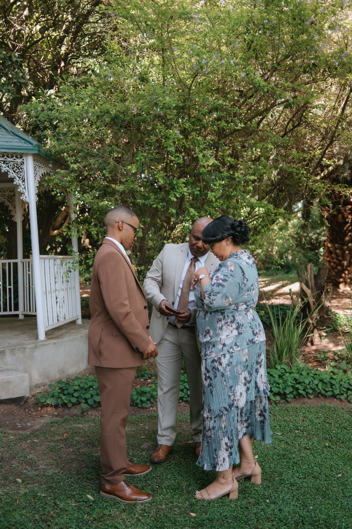 Three people share a prayer moment together in green garden near white lattice gazebo.